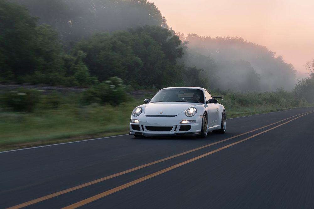 Weißer Porsche 911er auf Landstraße mit Sonnenaufgang und Tau im Hintergrund 