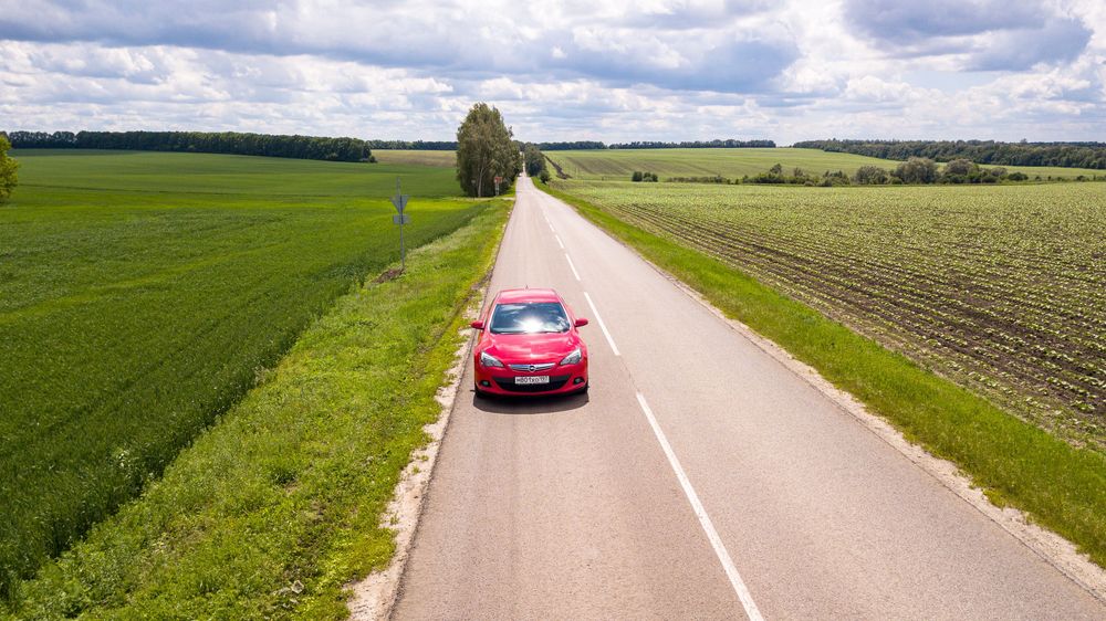 Roter Opel auf Landstraße zwischen zwei grünen Feldern bei sonniger und teils bewölkter Tageszeit