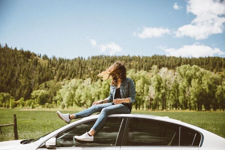 Glückliche Frau auf Auto vor sonnigem Wiese und Wald im Hintergrund