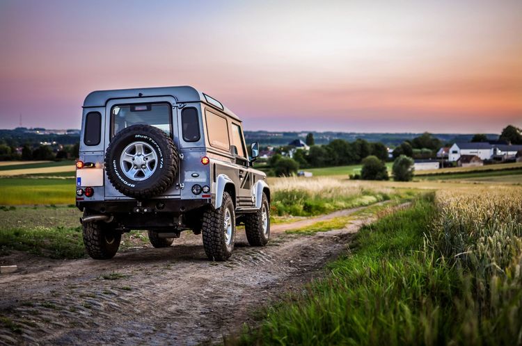 Grauer Land Rover Defender auf Feldweg beim Sonnenuntergang